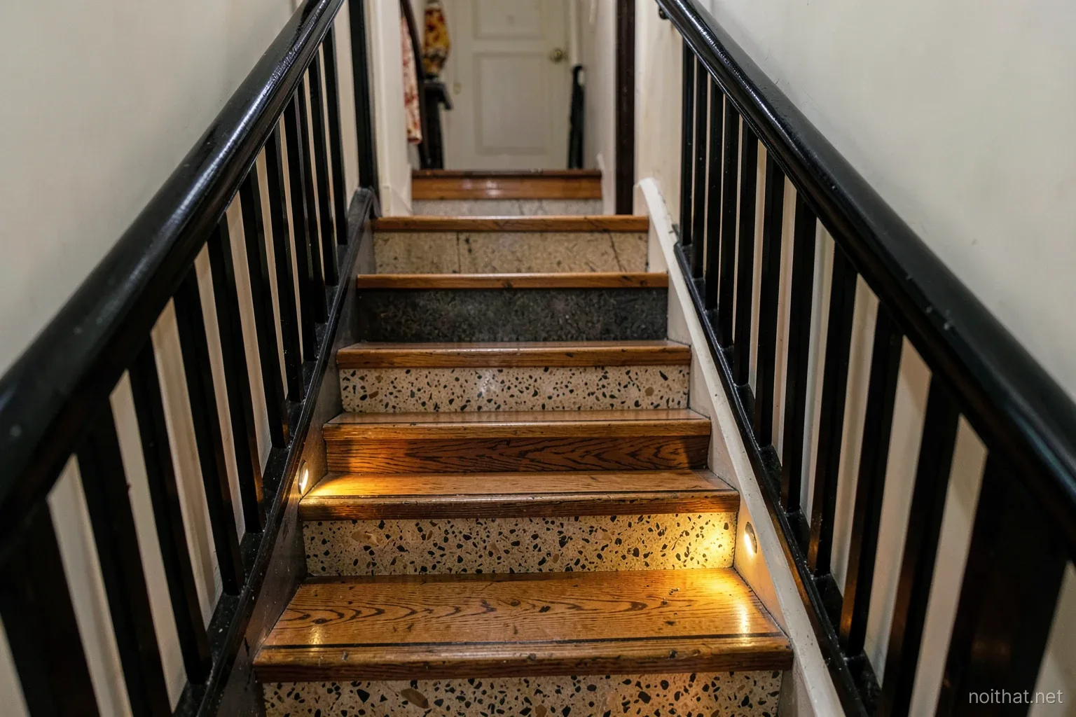 Staircase in a narrow Vietnamese townhouse after refresh, warm oak tread covers over old terrazzo steps, freshly painted black railing, small step lights glowing near the floor, view from below
