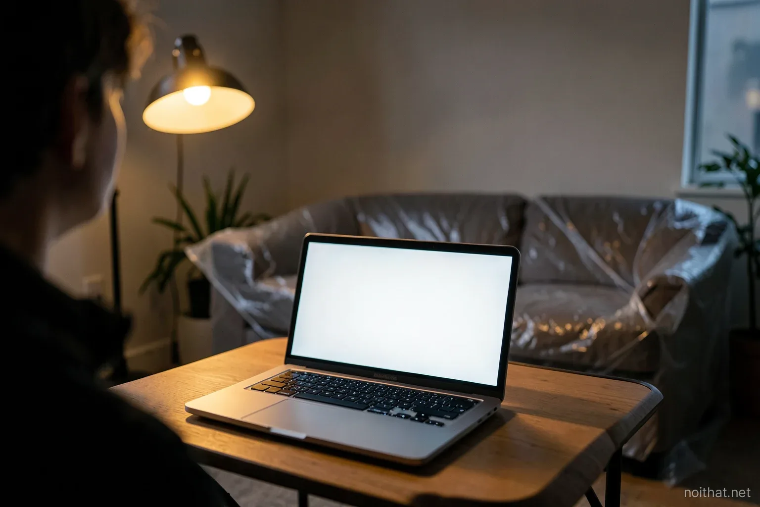 Laptop with a blank glowing screen on a folding table facing a half-finished living room where a sofa is still wrapped in plastic, dusk light mixed with a warm work lamp, over-the-shoulder view