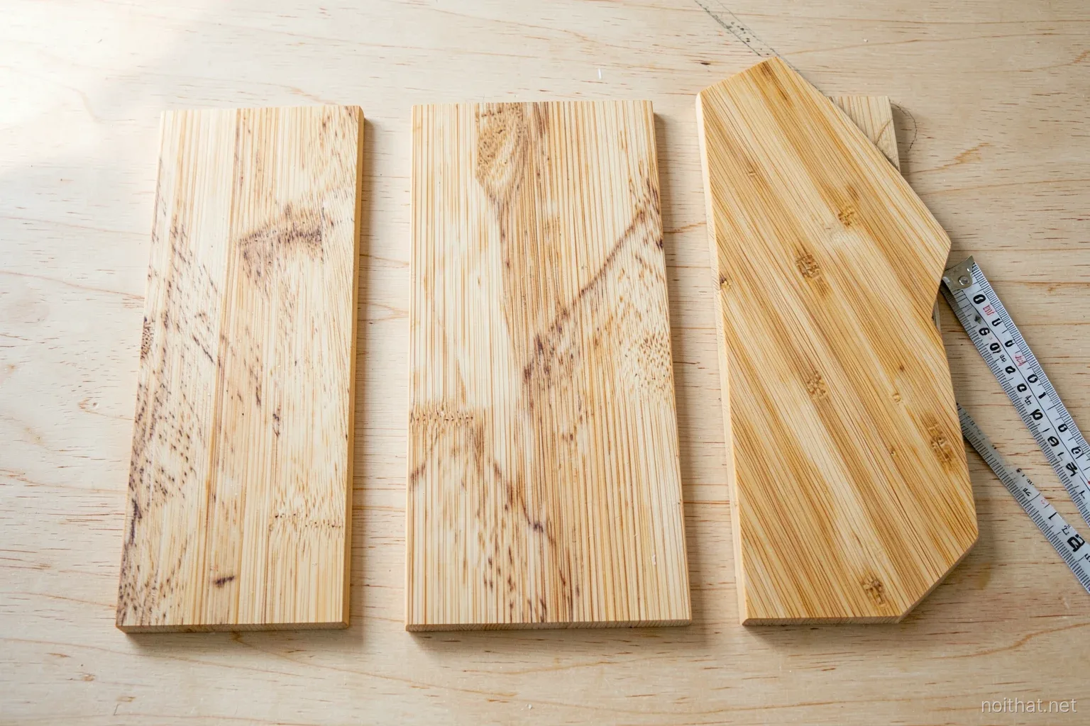 Top-down view of three bamboo board samples with fine parallel strips and a dense marbled strand pattern laid on a light plywood surface, morning window light, a tape measure nearby