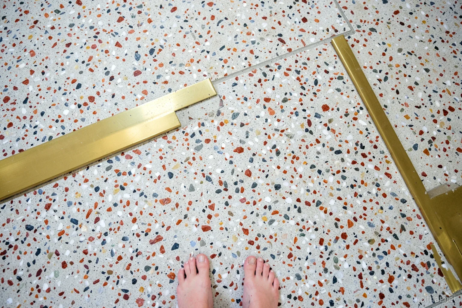 Overhead shot of a speckled terrazzo floor with white, grey and rust colored stone chips set in pale cement, a brass divider strip crossing the frame, bare feet at the edge, soft daylight